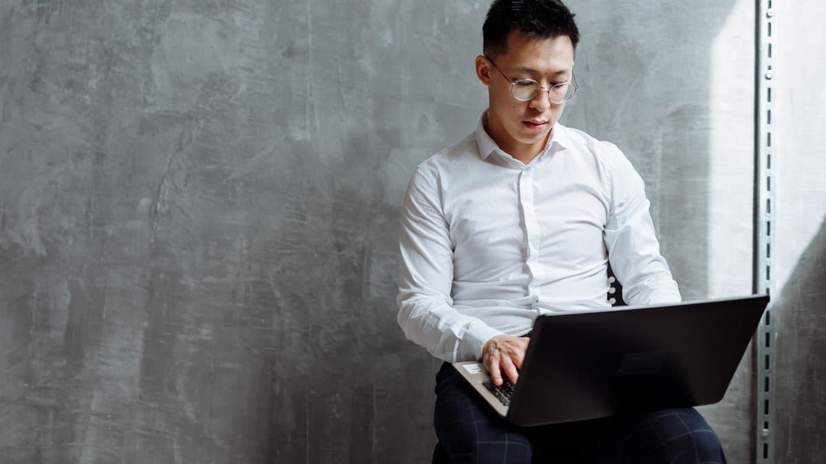 A man in a white shirt and glasses sits, focused on typing on a laptop, against a gray wall with a metal rack.