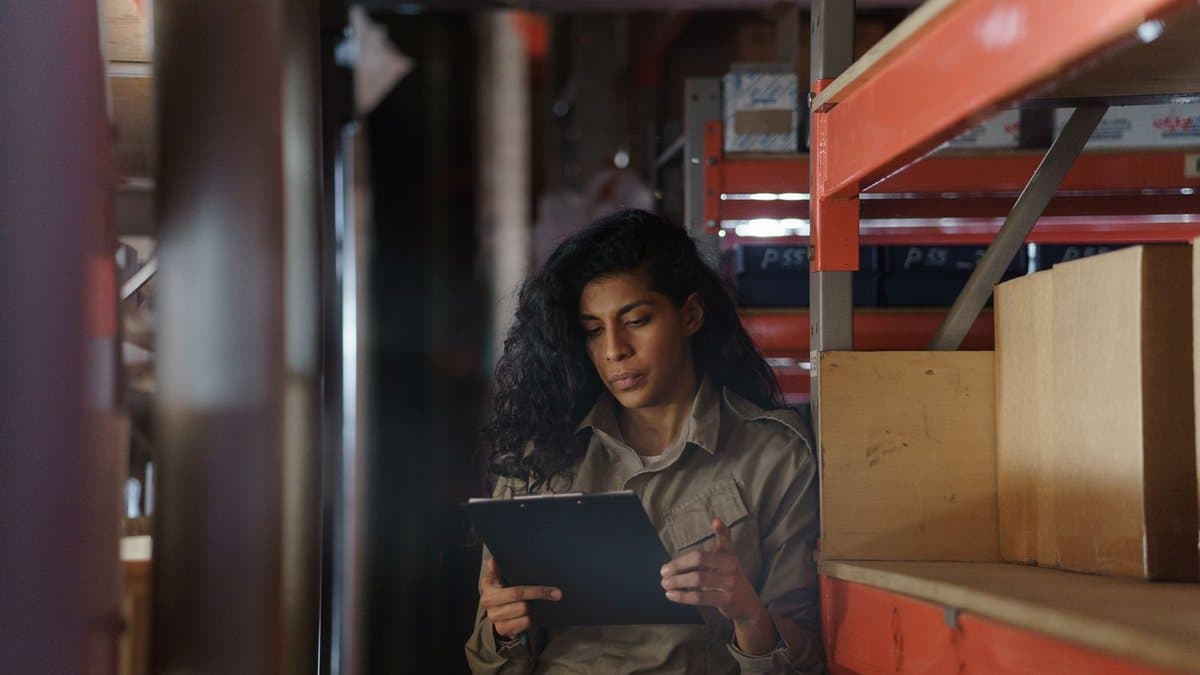 A woman with long, dark hair stands in a warehouse aisle, attentively examining a document on a clipboard.