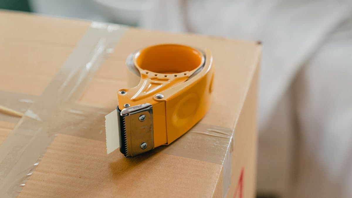 A yellow tape dispenser with exposed tape sits on a cardboard box, which is sealed with clear packing tape arranged in an X pattern.