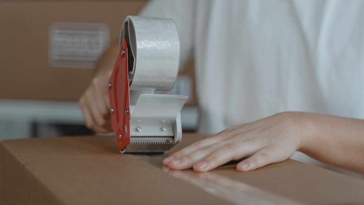 Hands use a red and white tape dispenser to seal a brown cardboard box with clear packing tape.