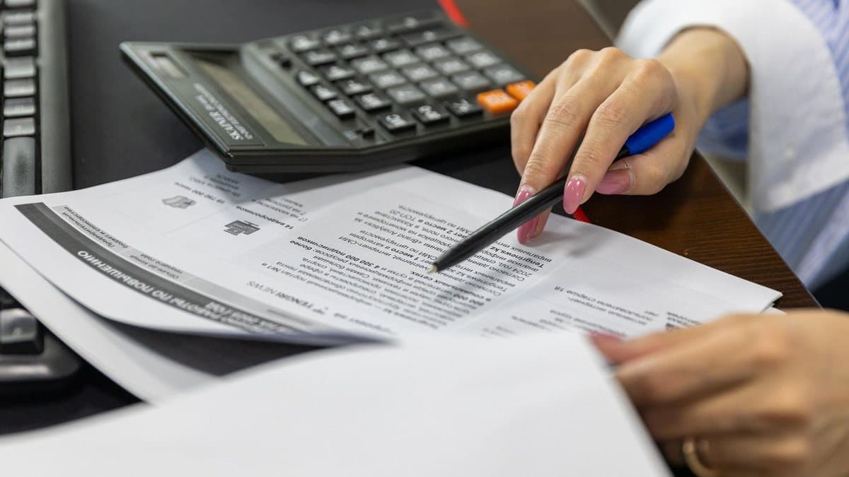 A person's hands are on a desk, one pointing with a pen at documents, with a calculator and a keyboard nearby.