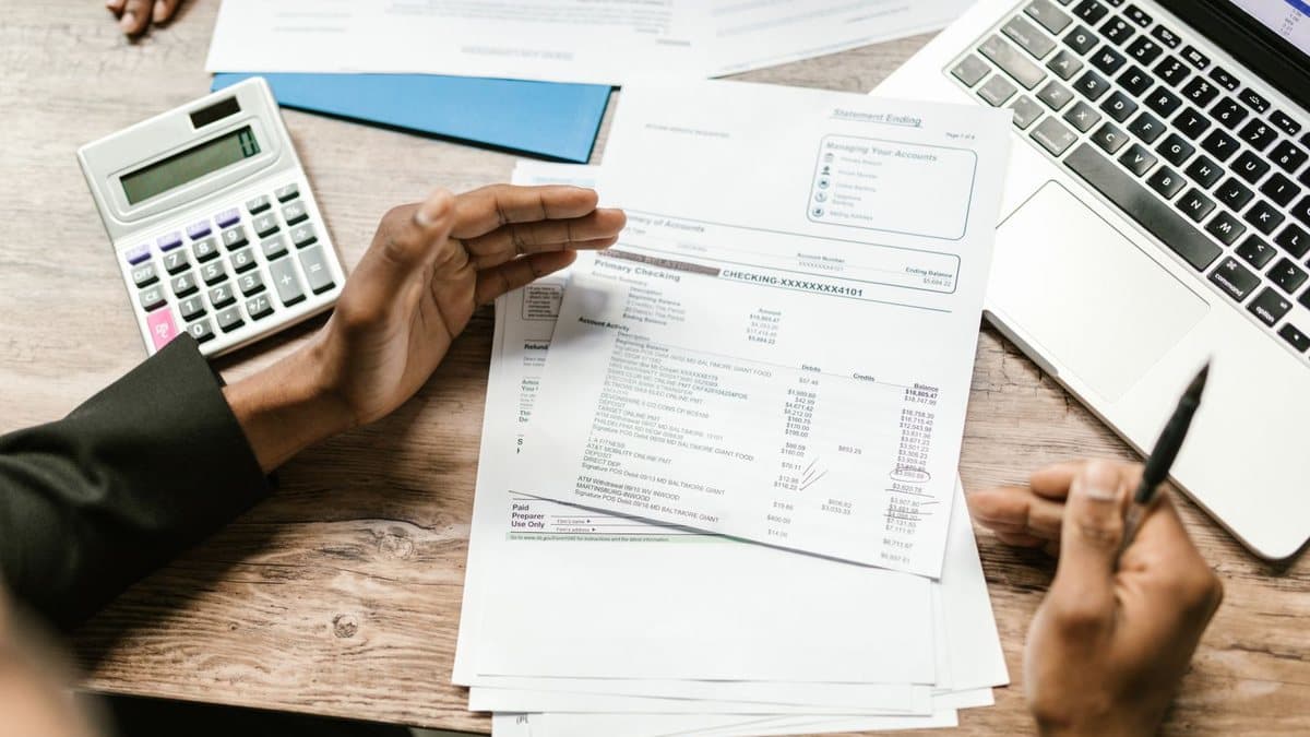 A person reviews financial statements on a wooden desk with a calculator and laptop, another hand visible.