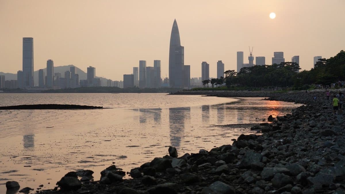 朦胧夕阳照亮了倒映在平静海湾中的现代城市天际线,人们在布满岩石的岸边行走。