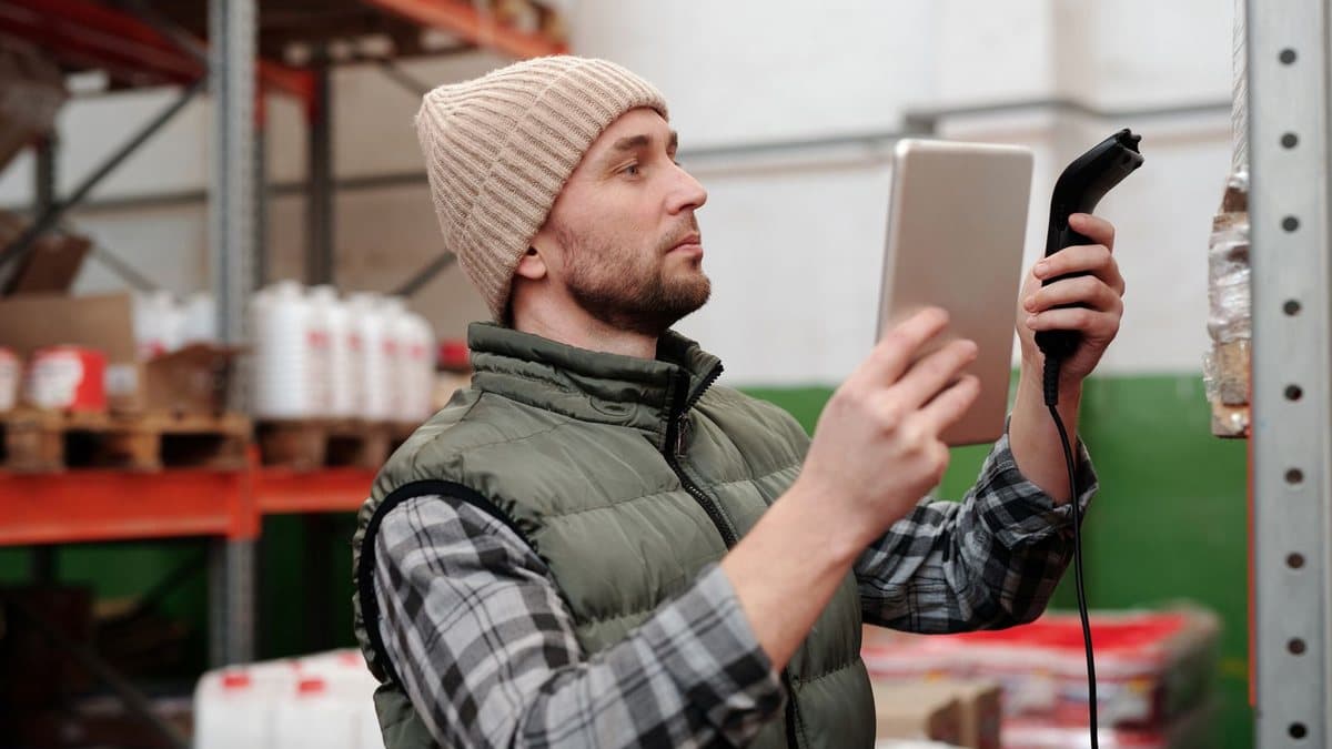 A man in a beanie and vest scans an item on a warehouse shelf using a barcode scanner and tablet.