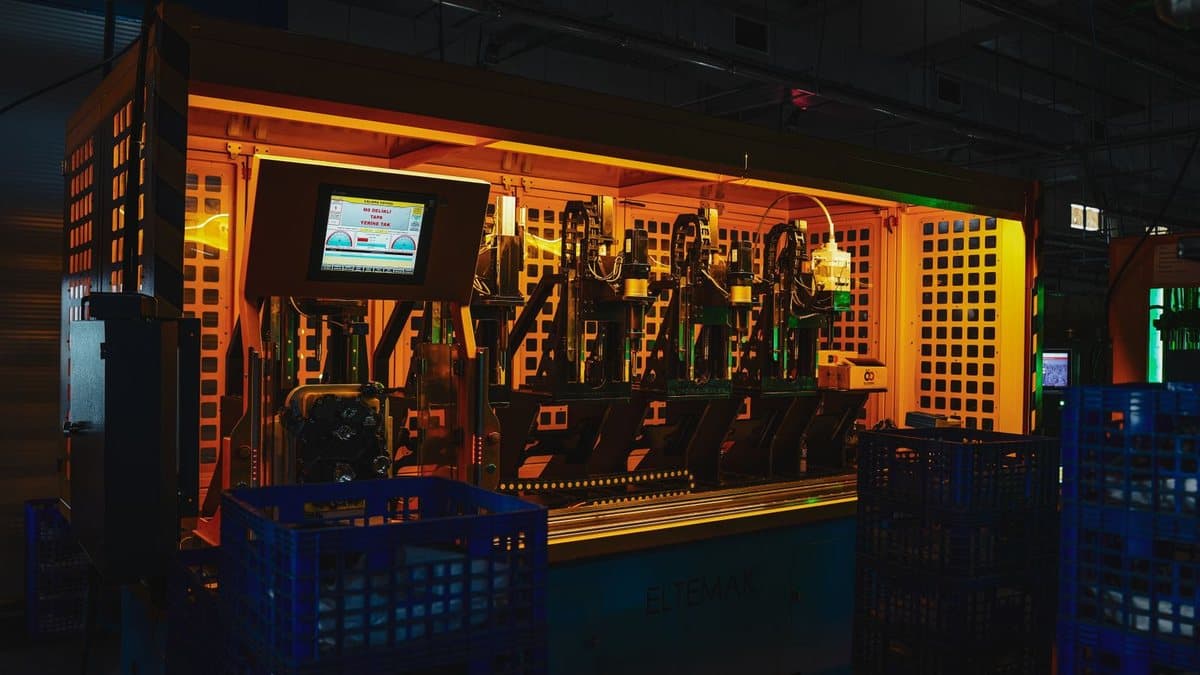 A large industrial machine glows orange in a dimly lit factory, surrounded by blue plastic crates and a digital control screen.