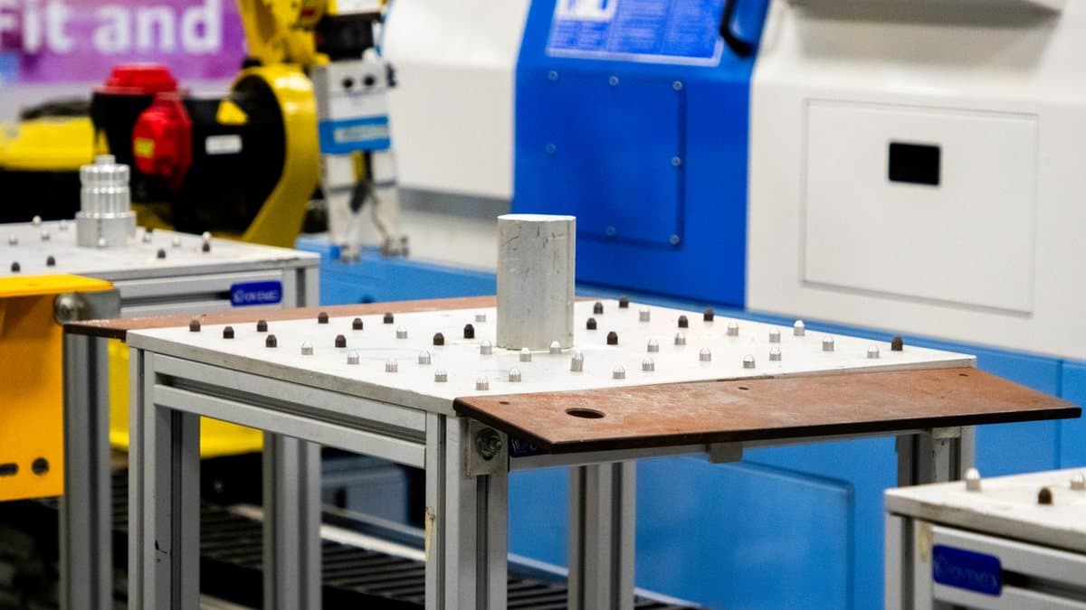 Several tables with grid-patterned pegs hold a cylindrical sample in a lab, with industrial machinery nearby.