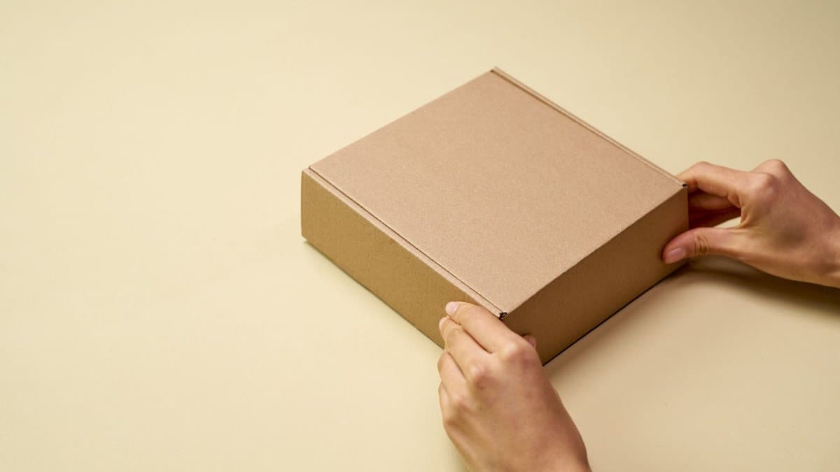 Two hands are gently closing a flat, brown cardboard box on a plain, light beige surface.