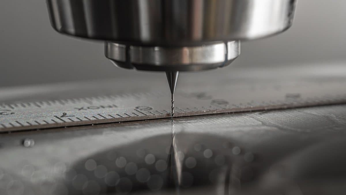 A tiny drill bit attached to a metallic spindle precisely touches a metal ruler with etched measurements.