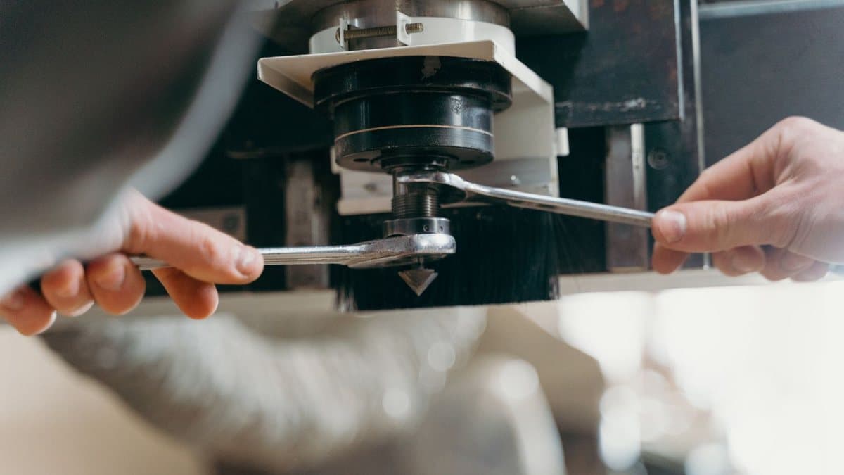 Two hands use wrenches to secure a pointed tool into a machine's spindle, with a dust brush around it.
