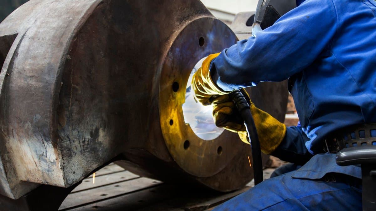 A welder in blue overalls and yellow gloves works on a large, dark metal component, creating bright sparks inside a circular opening.