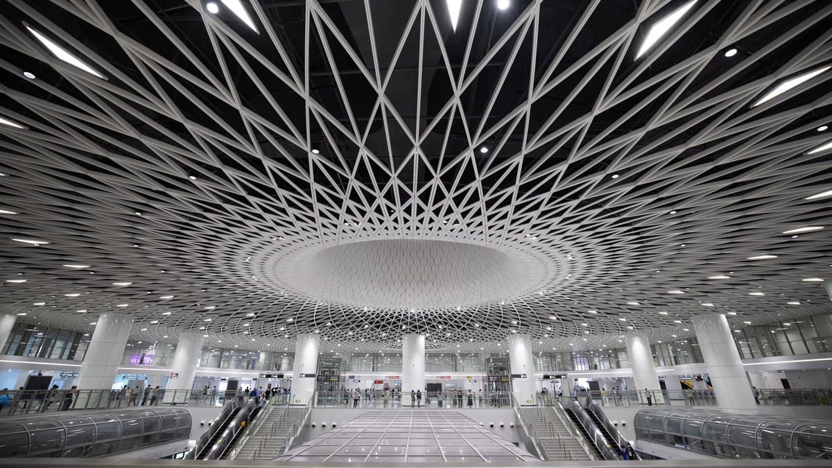 A vast transit hall under a white lattice ceiling that spirals into an oval at its centre, with escalators and stairs leading down and people walking along the upper level.
