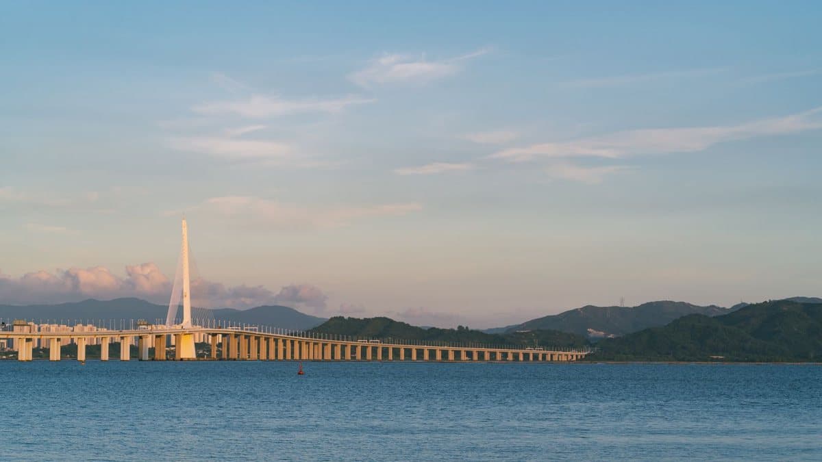 一座长长的斜拉桥横跨蓝色水面,远处是绿色的群山,天空呈现淡蓝与橙色。