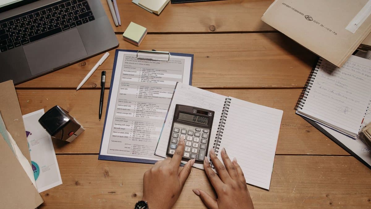 Hands operate a calculator on a cluttered wooden desk with a laptop, financial documents, notebooks, and pens.