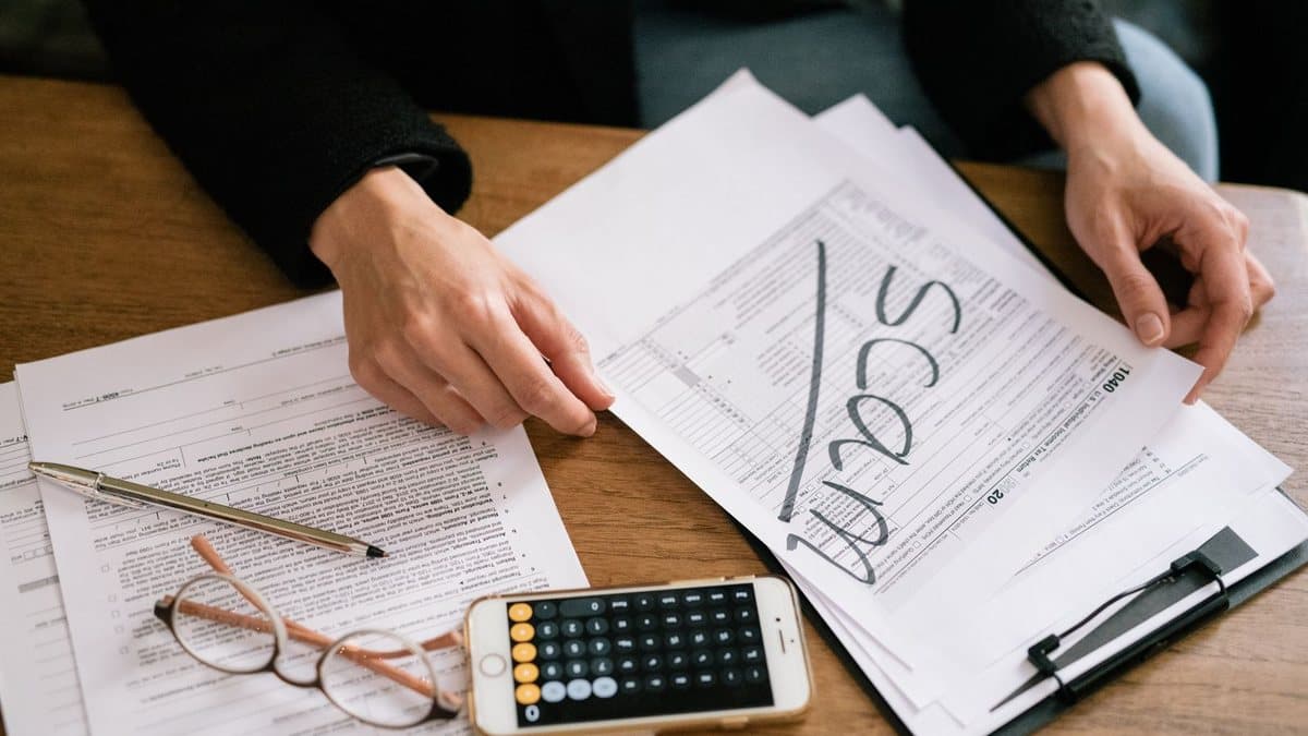 A person's hands are sorting through multiple tax forms on a wooden desk, one marked "SCAM", beside a pen, eyeglasses, and a smartphone calculator.