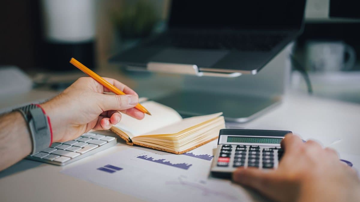 Hands on a desk hold a pencil over a notebook, while another hand operates a calculator amidst work documents and a laptop.