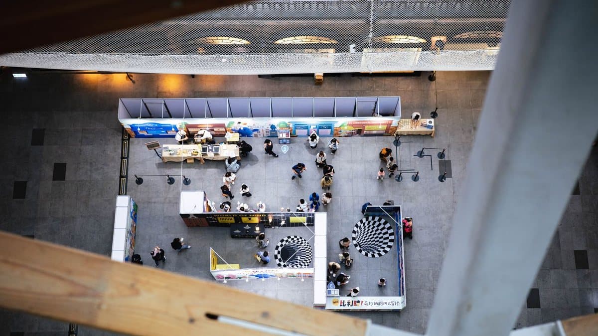 An overhead view shows an indoor exhibition with many people browsing various booths and two large optical illusion floor patterns.