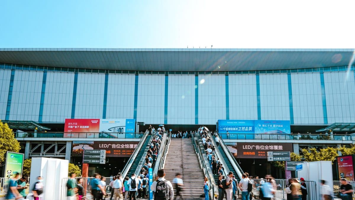 Crowds of people move up escalators and a staircase towards a large building with advertisements under a clear blue sky.
