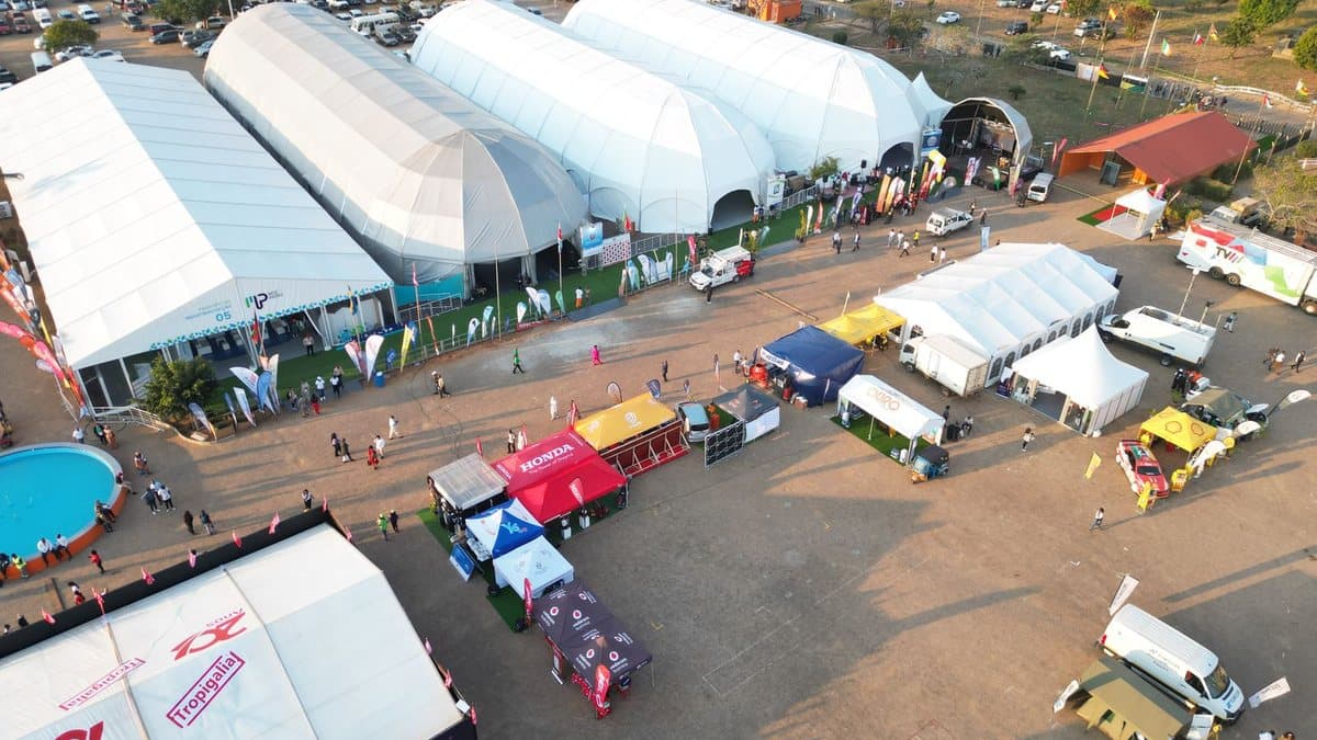 An outdoor exhibition features large white tents and various smaller vendor booths with many people and vehicles on a dirt lot.