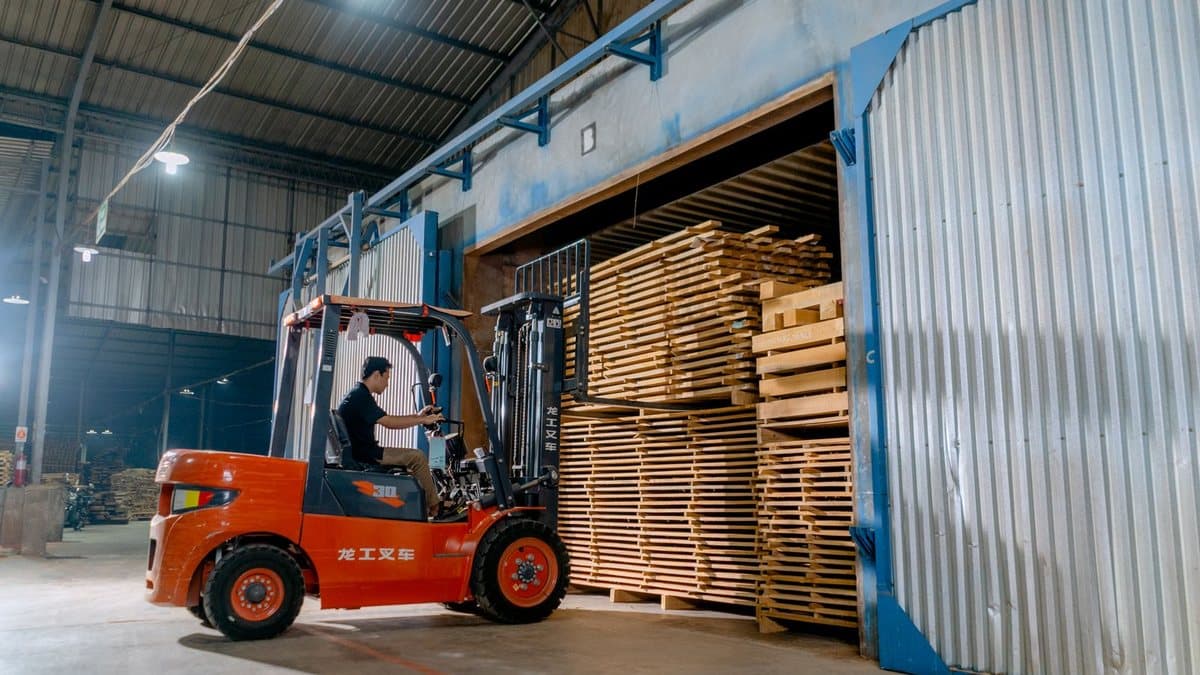一名男子驾驶橙色叉车,将大量浅色木板堆叠送入波纹金属工业烘干机或储存单元。