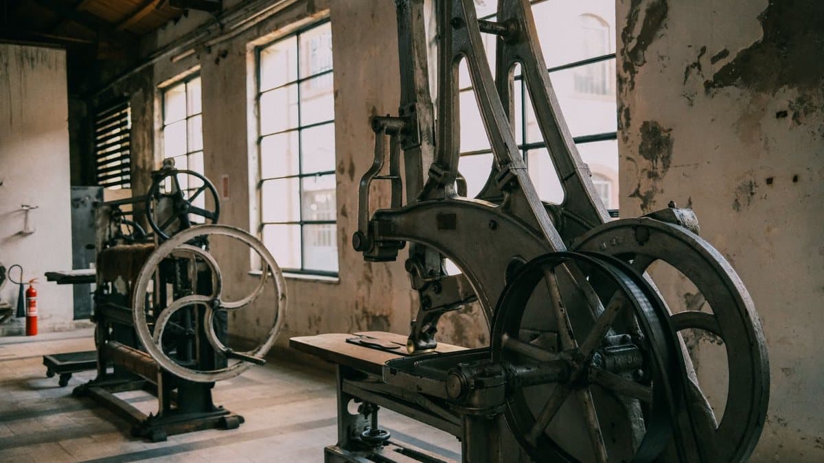 Two large, old industrial machines with prominent wheels stand in a derelict factory hall with tall windows and peeling walls.