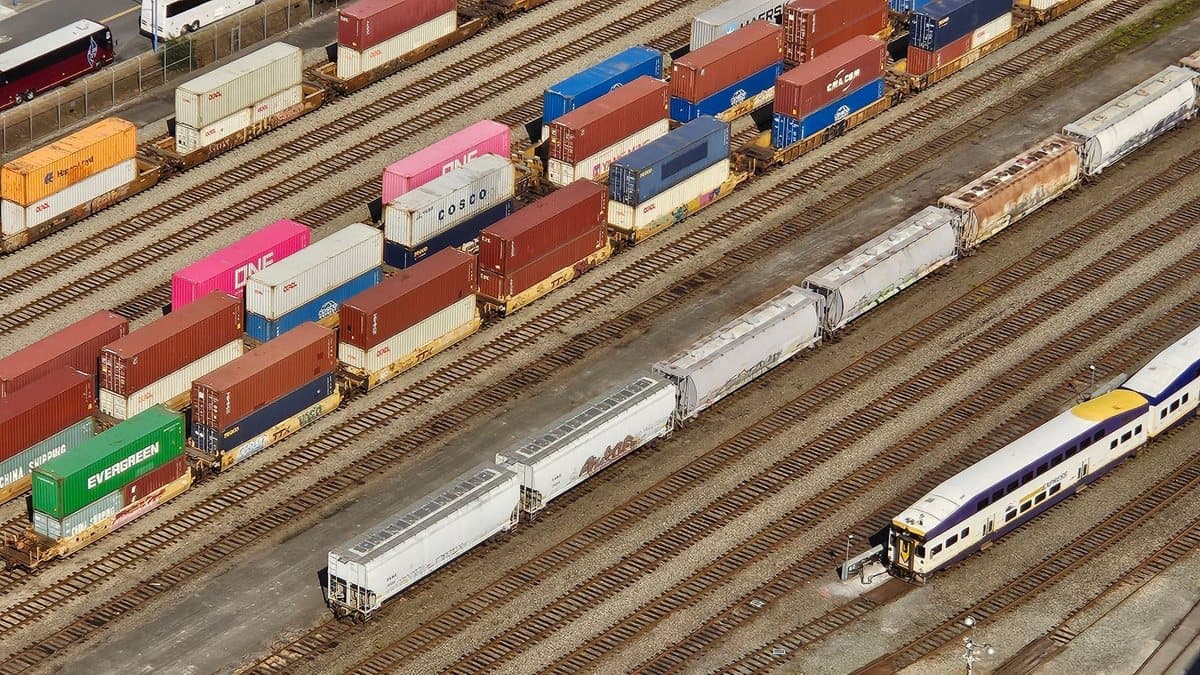 Multiple parallel rail tracks fill a train yard, where freight trains with colorful stacked containers and a parked passenger train are visible.