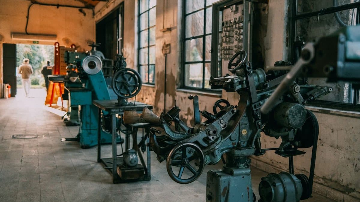 Vintage manufacturing machines line a rustic workshop, with two people walking towards a bright outdoor exit.