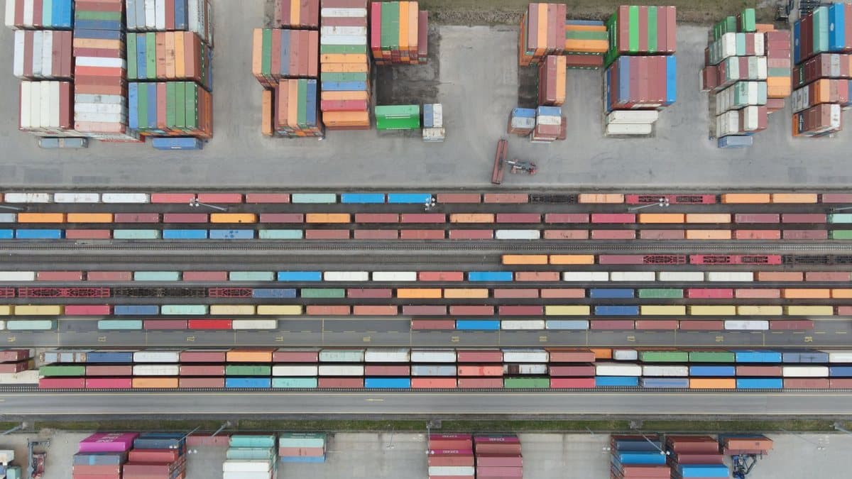 An overhead view shows colorful shipping containers stacked and on railcars in a vast intermodal yard, with a small vehicle.