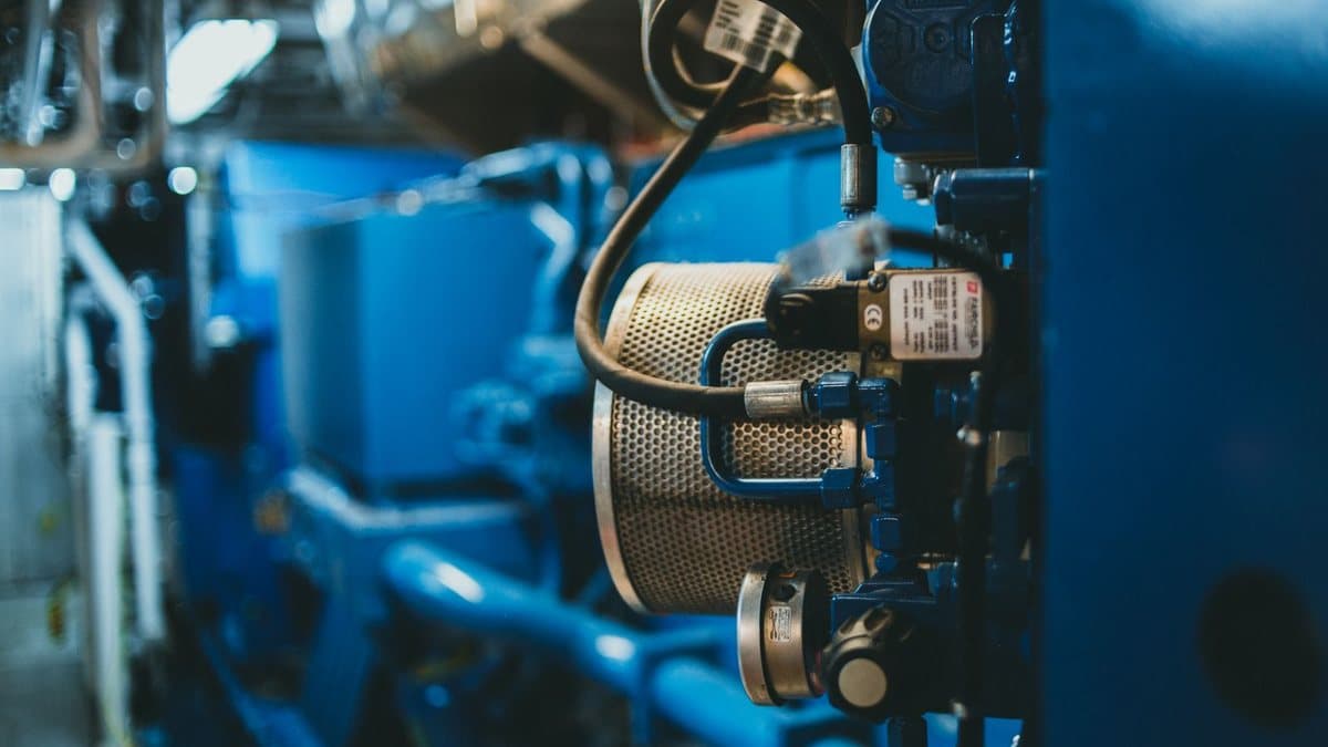 Close-up of a blue industrial machine, featuring a perforated cylindrical component, various pipes, hoses, and gauges.