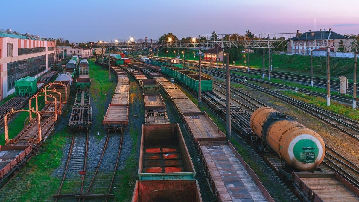 Numerous freight trains and tracks fill a busy railway yard at dusk, flanked by industrial buildings and distant trees.