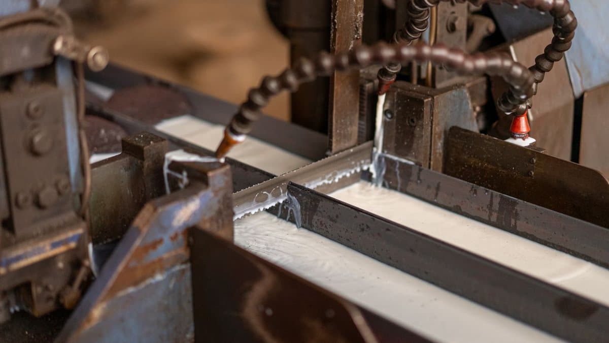 An industrial band saw cuts a metal piece, with milky coolant flowing from two hoses onto the blade and workpiece.