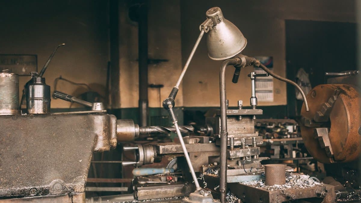 An old, oil-stained metal lathe, topped with an adjustable lamp, is covered in metal shavings within a dimly lit workshop.